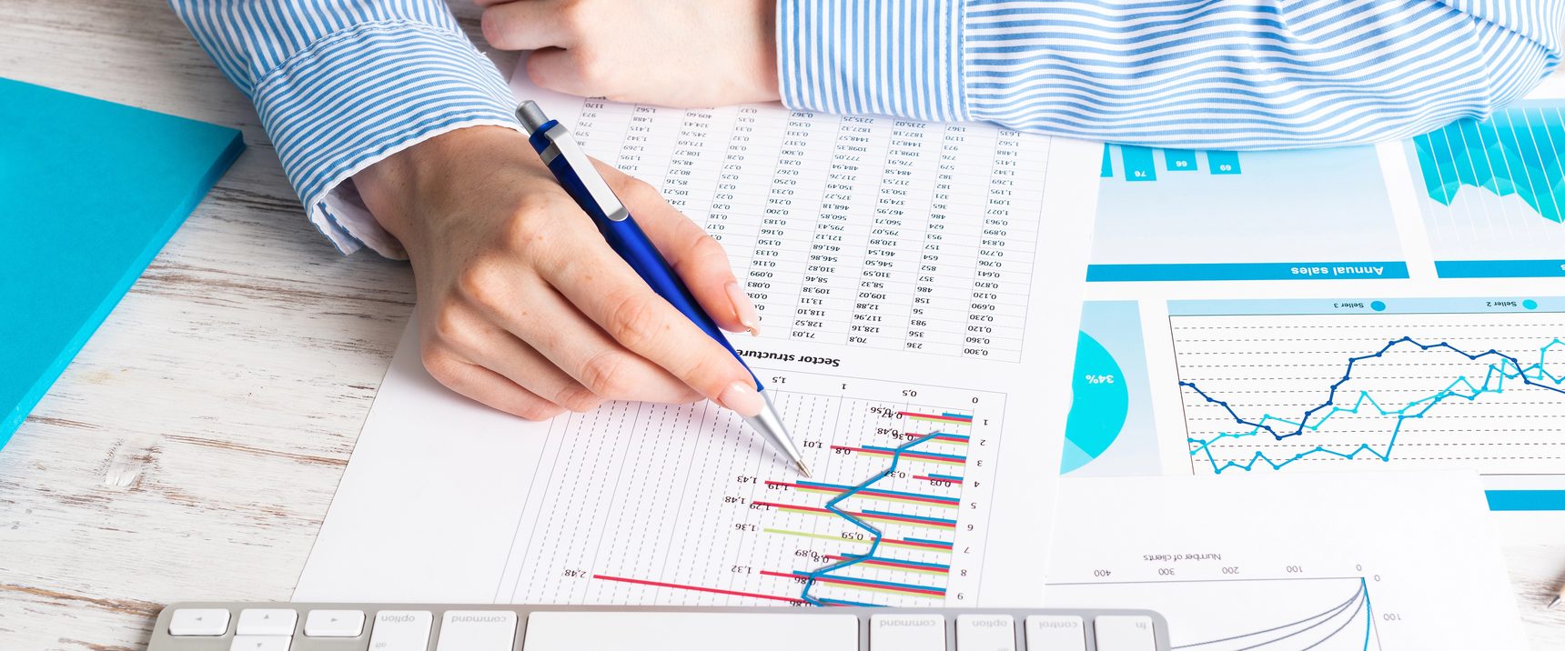 A hand holding a pen over a printed spreadsheet and a set of charts, beside a keyboard